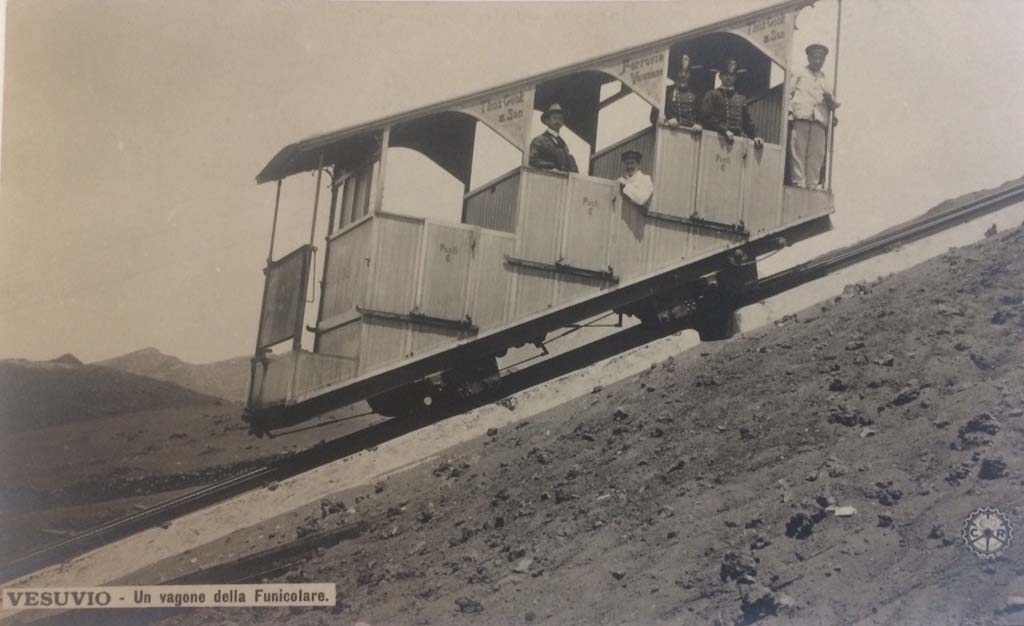 Postcard showing a Wagon of the Vesuvius Funicular, with handwritten note on rear-  “The 1904 Funicular only lasted 2 years”.  
Photo courtesy of Rick Bauer.
This photo must be dated between 1904 when the cars were introduced and 1906 when they were destroyed..
The funicular was modernised at the same time as Cook’s Pugliano to Vesuvius light railway was opened in 1904.
The monorails had been removed and the track was now a one metre gauge single track with a central passing loop.
These cars were new in 1904 and seated 18 with 6 standing. 
Thomas Cook and Son’s name is on the upper part of the structure. The wording on each of the three doors is “Posti 6” (6 places).
Two years later, in a tremendous eruption, on 7 and 8 April 1906 the lower and upper station, the equipment, the machinery, the two funicular cars were destroyed.
Everything was buried under a 20-30m high ash blanket.
Although the light railway as far as the hotel Eremo was quickly dug out, the funicular had to wait. 
Only in 1909, according to the project of the engineer Enrico Treiber, were the works for a new funicular ended and it reopened in 1910.
