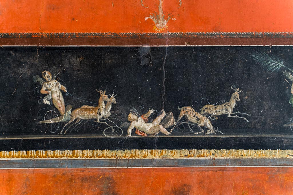 VI.15.1 Pompeii. March 2023. 
Lower centre of east wall, detail of part of painted panel with cupids in a chariot race. Photo courtesy of Johannes Eber.
